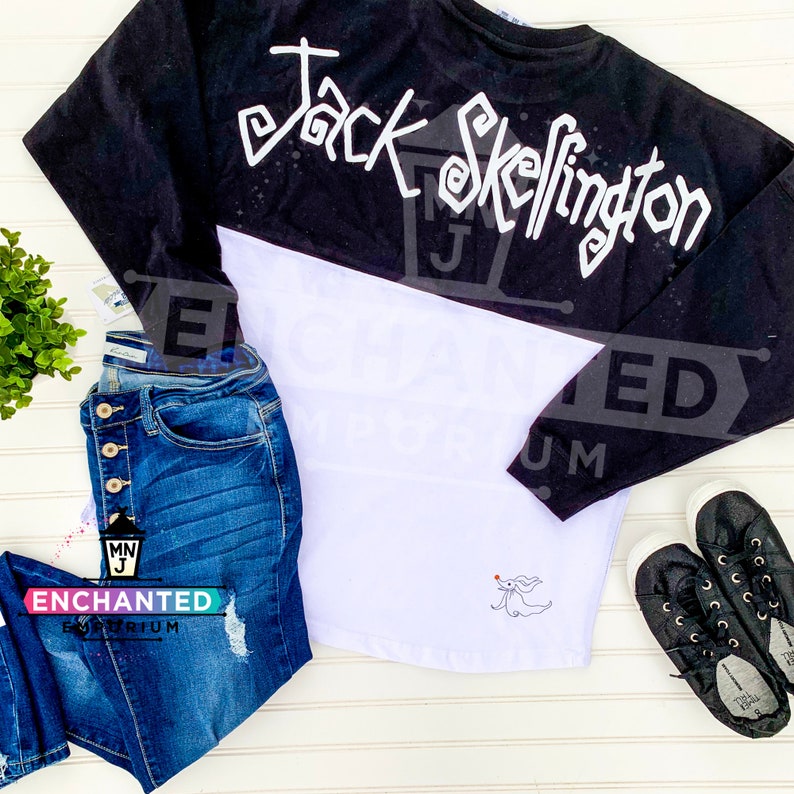 May include: Black and white long-sleeved shirt with a white graphic of Jack Skellington's name on the back and a white graphic of Zero on the front. The shirt is paired with blue jeans.