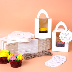Puede incluir: Cajas blancas para cupcakes con ventanas transparentes, una pila de cajas en blanco, cuerda marrón y etiquetas de regalo para cupcakes que dicen "hecho a mano con amor". Las cajas tienen un asa y son perfectas para empaquetar cupcakes.