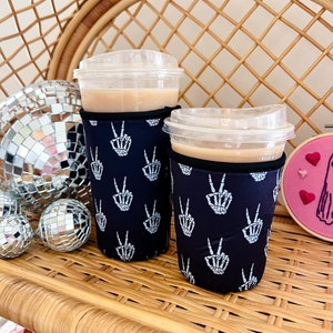May include: Two black and white coffee cup sleeves with a skeleton hand peace sign pattern. The sleeves are on two clear plastic cups with brown liquid inside.