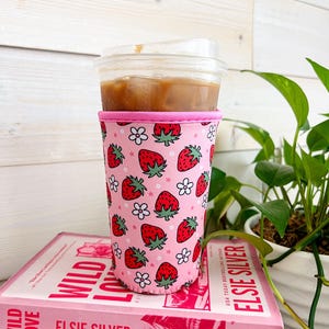May include: A pink and white insulated cup sleeve with a strawberry and flower pattern. The sleeve is on top of two books, one titled "Wild Love Elsie Silver" and the other titled "People We Meet on Vacation".