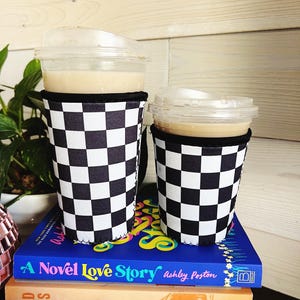 May include: Two iced coffee drinks in clear plastic cups with lids, each in a black and white checkered cup sleeve. The drinks sit on a stack of books, including "A Novel Love Story" by Ashley Poston.