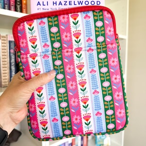 May include: A rectangular fabric sleeve with a red zipper, featuring a vertical striped pattern of pink, green, and white with stylized floral motifs. A book by Ali Hazelwood is tucked inside the sleeve, showing the author's name in blue text.