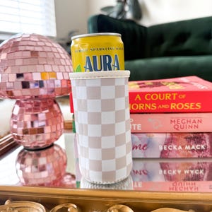 May include: A yellow can of "AURA" herbal sparkling water sits in a beige and white checkered can cooler. A pink disco ball lamp and a stack of books are in the background. The books include "A COURT OF THORNS AND ROSES" by Meghan Quinn.