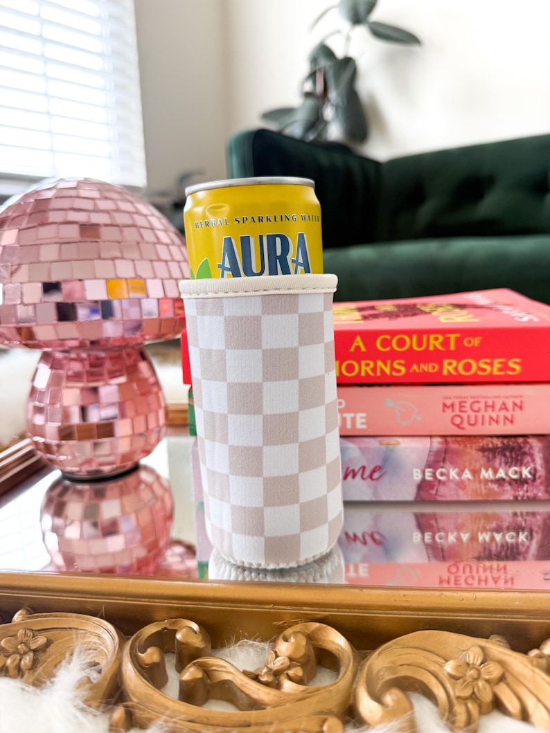 May include: A yellow can of "AURA" herbal sparkling water sits in a beige and white checkered can cooler. A pink disco ball lamp and a stack of books are in the background. The books include "A COURT OF THORNS AND ROSES" by Meghan Quinn.
