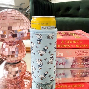 May include: A light blue beverage can cooler with a pattern of white geese wearing blue bows. A yellow can of sparkling water is inside the cooler. The cooler is on a gold tray with a stack of books and a pink disco ball.