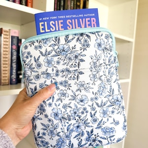 May include: A rectangular protective sleeve featuring a white background with a blue floral pattern and a light blue zipper closure. A book with a blue cover and the text #1 NEW YORK TIMES BESTSELLING AUTHOR ELSIE SILVER is tucked inside.