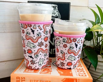 May include: Two pink and white Halloween-themed coffee cup sleeves with a design of ghosts, skulls, candy corn, and bats. The sleeves are on top of a stack of books with the titles "Wild Eyes", "Happy Place", and "A Novel Love Story".