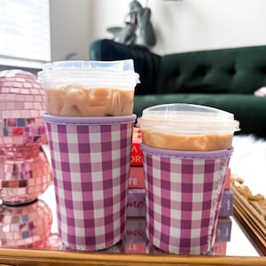 May include: Two iced coffee drinks in clear plastic cups with lids, resting in purple and white gingham patterned cup sleeves. The drinks sit on a gold tray with decorative edges, with a pink disco ball in the background.