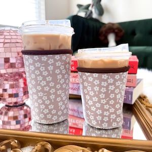 May include: Two iced coffee drinks in clear plastic cups with lids, each in a floral patterned sleeve. The sleeves are light pink with white flowers and a brown trim. The drinks sit on a gold-framed mirror.
