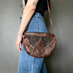 Può includere: Una borsa a tracolla in pelle marrone a forma di mezza luna. La borsa presenta un pannello centrale strutturato marrone scuro e un design a pieghe nella metà inferiore. Il cinturino è di un sottile colore marrone abbinato.