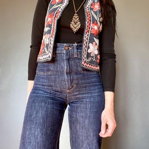 Puede incluir: Un chaleco negro sin mangas con bordados florales en rojo y rosa vibrantes y ribete en zigzag blanco, usado sobre una camiseta negra de cuello alto y manga larga, combinado con jeans azul oscuro de tiro alto y un collar con colgante de filigrana dorada.