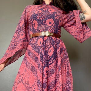 Puede incluir: Vestido fluido de estilo vintage en tonos rosa y morado, con estampado floral. El vestido tiene mangas largas y acampanadas y se ciñe a la cintura con un cinturón marrón con hebilla dorada. El vestido tiene un cuello alto.