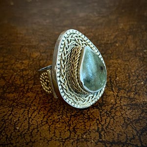 Può includere: Un anello d'argento con una grande pietra verde a forma di goccia. L'anello presenta un'intricata lavorazione in metallo color oro attorno alla pietra e sulla fascia. L'anello è posto su uno sfondo marrone.