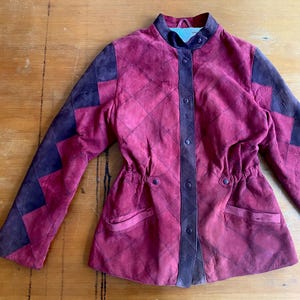 May include: A burgundy suede jacket with a quilted diamond pattern and dark purple accents. The jacket features a stand-up collar, button closures, and a drawstring waist. The sleeves have a geometric design.