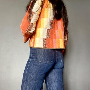 Peut inclure: Une veste matelassée présentant un motif patchwork de panneaux rectangulaires en tissu orange, jaune et crème avec des motifs floraux, associée à un jean bleu foncé. La veste a des manches longues et un ourlet droit avec une bordure marron.