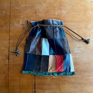 Può includere: Una borsa a coulisse in pelle patchwork in vari colori tra cui nero, marrone, rosso e bianco. La borsa ha una frangia verde sul fondo e un cordoncino marrone con estremità decorative. La borsa è su una superficie di legno.