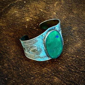 May include: A silver cuff bracelet with an oval, green gemstone. The bracelet has an ornate, etched design with a turquoise patina. The gemstone has a blue accent. The cuff is open and adjustable.