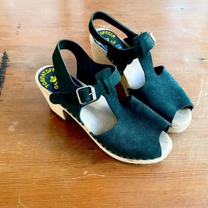 Pode incluir: Um par de tamancos de camurça verde-escura com biqueira aberta e solas de madeira. Os sapatos apresentam um design de tira em T com uma fivela de metal prateado ajustável no tornozelo. As palmilhas internas trazem o texto Torpatoff e Offeln.