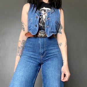 Può includere: Un gilet di jeans con bottoni e una cintura in pelle marrone con fibbia decorativa. Il gilet è indossato sopra una maglietta nera con grafica e jeans blu a vita alta. L'outfit ha un'estetica vintage e retrò.