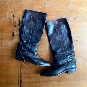 Könnte beinhalten: Ein Paar schwarze kniehohe Lederstiefel mit Schlangenmuster am oberen Schaft. Die Stiefel haben eine spitze Zehenpartie und einen kleinen Absatz. Die Stiefel sind auf einer Holzoberfläche ausgestellt.