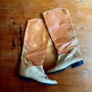Könnte beinhalten: Ein Paar kniehohe Stiefel in den Farben Beige und Creme. Die Stiefel haben ein dekoratives Muster an der Seite und einen niedrigen Absatz. Die Stiefel sind aus Leder und haben einen klassischen Western-Stil.