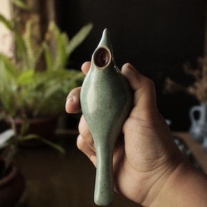 Op de afbeelding: Een groene keramische neti pot in de vorm van een vogel met een klein gaatje bovenaan. De pot is gevlekt met bruin en heeft een glad, rond lichaam.