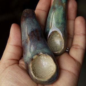 May include: Two handcrafted ceramic pieces with a teardrop shape, held in a hand. The pieces have a glossy finish with shades of blue, green, and brown. Each piece features a textured, light-colored interior.