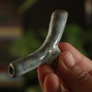 May include: A Y-shaped ceramic pipe with a smooth, glossy finish in shades of mottled blue, grey, and brown. The object features a hollow cylindrical stem and a small bowl section, showcasing a handcrafted, organic form with a glazed surface.