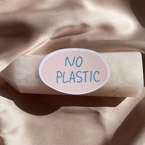 May include: A light-colored crystal with an oval sticker that reads "NO PLASTIC" in blue text. The crystal is resting on a satin fabric.