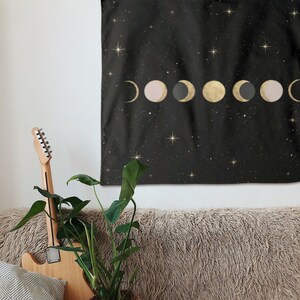 May include: A black tapestry with gold and pink moons and stars on a black background hanging on a white wall above a brown fuzzy couch with a potted plant and a guitar leaning against it.