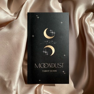 Puede incluir: Un libro de guía de tarot negro con un diseño en lámina dorada de dos lunas crecientes y estrellas. El título "MOONDUST TAROT GUIDE" está impreso en lámina dorada en la portada.