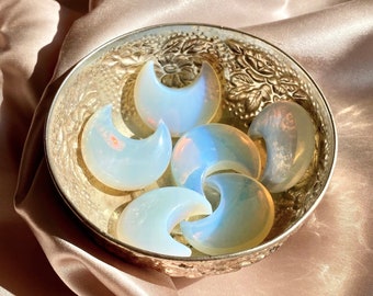 May include: A bowl filled with six iridescent opalite crescent moon shaped stones. The bowl is silver and has an ornate design.