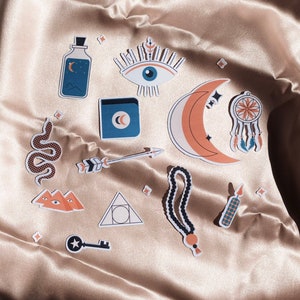 May include: A set of 12 colorful stickers featuring various mystical and spiritual symbols, including an eye, a crescent moon, a snake, an arrow, a dreamcatcher, a key, a bottle, a triangle, a candle, and a string of beads.