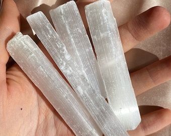May include: Four white selenite wands, also known as selenite sticks, are arranged in a pile. The wands are smooth and have a natural, striated texture.
