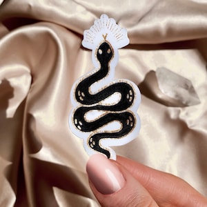 May include: A white sticker with a black snake design. The snake is coiled and has a crown on its head. The sticker has a gold outline.