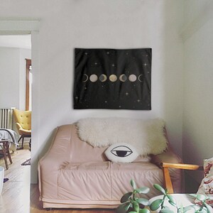 May include: A black tapestry with a gold and white moon phases design on a black background with white stars. The tapestry is hanging on a white wall above a pink couch with a white fur throw and a black and white eye pillow.