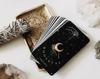 May include: A deck of "MOONDUST" tarot cards with a black background and gold crescent moon design. The cards rest on a gold-colored tray with a sun and moon design, next to a bundle of sage and crystals.