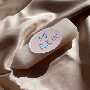 May include: A light-colored, pointed crystal with a sticker that reads "NO PLASTIC" in blue text. The sticker is oval-shaped with a light pink background. The crystal is resting on a satin fabric.