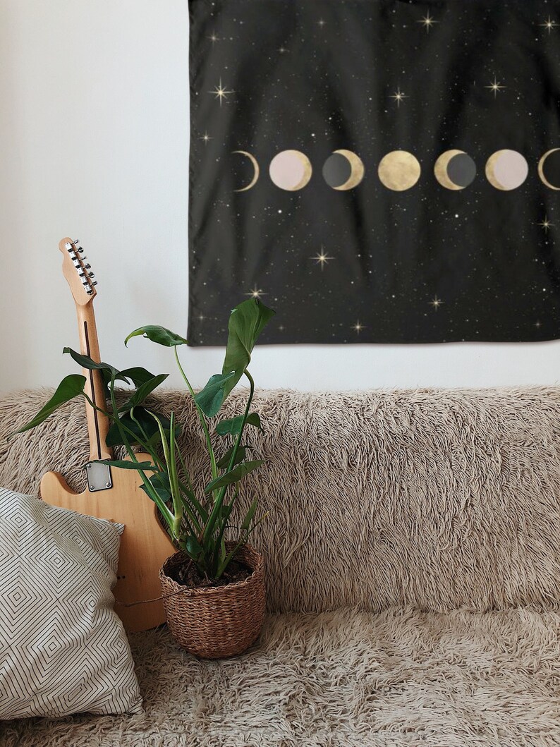 May include: A black tapestry with gold and pink moons and stars on a black background hanging on a white wall above a brown fuzzy couch with a potted plant and a guitar leaning against it.