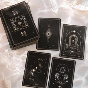 May include: A deck of black tarot cards with gold celestial illustrations and text including Dreamspell Tarot, The Star, Eight of Wands, Five of Wands, and Two of Pentacles. The cards feature intricate line art of hands, stars, and moons.
