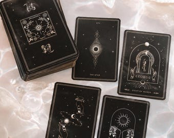 Może przedstawiać: Talia czarnych kart tarota ze złotymi niebiańskimi ilustracjami i napisami, w tym Dreamspell Tarot, The Star, Eight of Wands, Five of Wands oraz Two of Pentacles. Karty zawierają szczegółowe rysunki liniowe przedstawiające dłonie, gwiazdy i księżyce.
