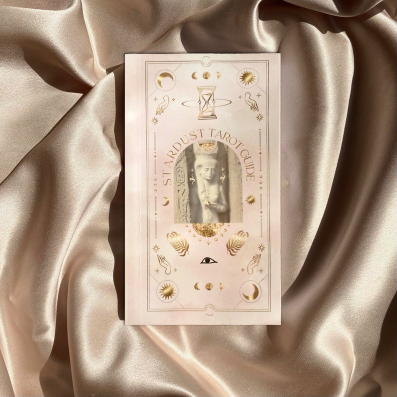 May include: A tarot guide titled "Stardust Tarot Guide" with a light pink cover. The cover features gold celestial illustrations, including moons, stars, and an hourglass. The center image is a statue. The guide is on a satin surface.