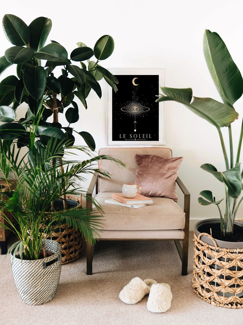 May include: A beige armchair with a pink pillow and a stack of books. A white mug sits on top of the books. A black and gold framed print with the text "LE SOLEIL" hangs on the wall behind the chair. Two potted plants, one with green leaves and the other with large, green leaves, are on either side of the chair. Two white fluffy slippers are on the floor in front of the chair.