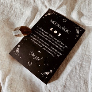 Puede incluir: Un libro negro con el t&iacute;tulo "Moon Magic" y el texto "Behold the wonder of the Dreamspell Tarot. This eye-catching guidebook is a dreamscape of magic, mysticism, and divine feminine energy. Created with intention in our NYC design studio, each page is brimming with wisdom that will help guide you on your tarot journey." El libro est&aacute; sobre una superficie blanca.