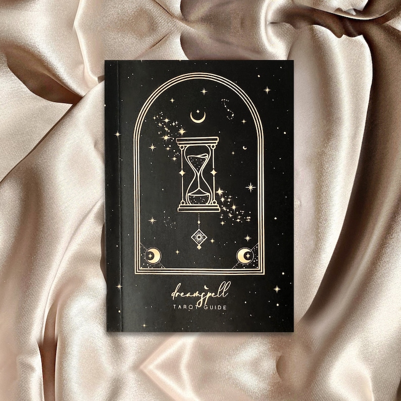 Puede incluir: Un libro negro con una ilustraci&oacute;n dorada de un reloj de arena, estrellas y lunas. El t&iacute;tulo del libro es "dreamspell TARO GUIDE".