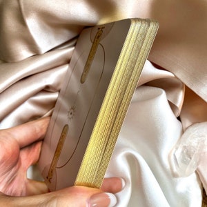 May include: A deck of tarot cards with gold gilded edges. The cards have a light beige background with gold line art and symbols. The deck is held in a hand against a backdrop of satin fabric.
