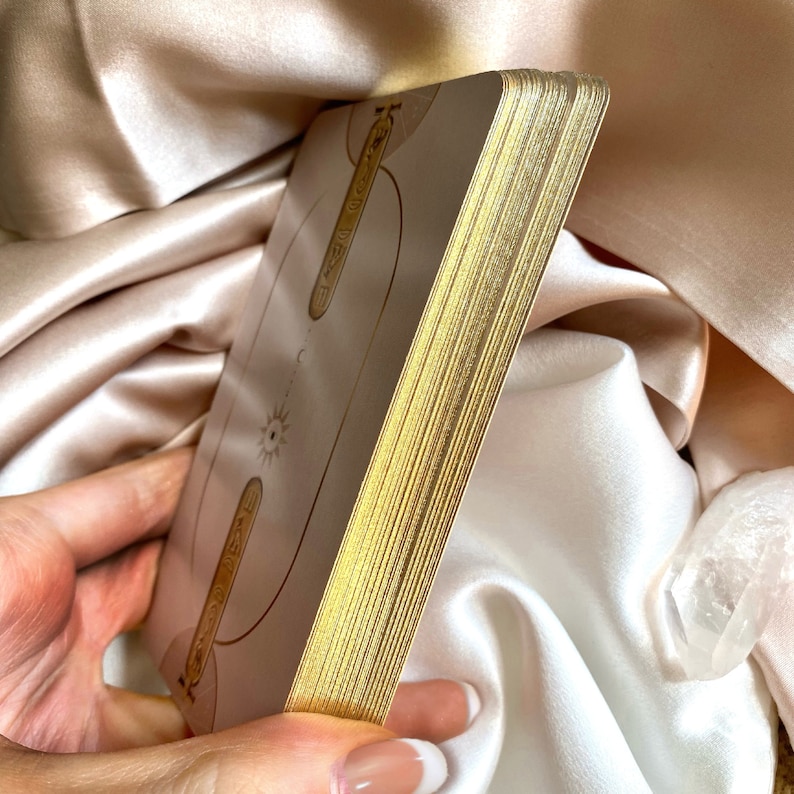 May include: A deck of tarot cards with gold gilded edges. The cards have a light beige background with gold line art and symbols. The deck is held in a hand against a backdrop of satin fabric.