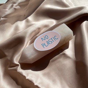 May include: A light-colored crystal point with a sticker that reads "NO PLASTIC" in blue text on a pink oval. The crystal is set against a backdrop of a silky, beige fabric.