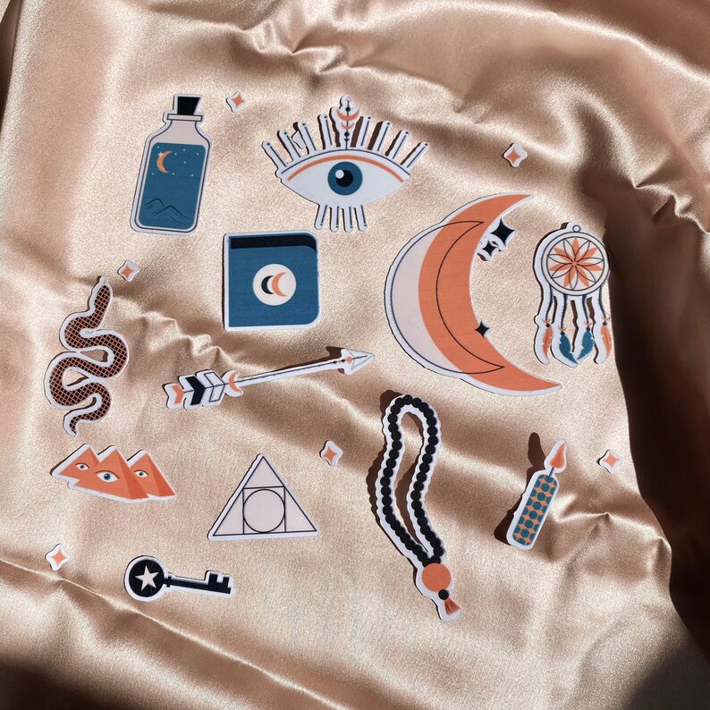 May include: A set of 10 colorful stickers featuring various mystical and spiritual symbols, including an eye, a crescent moon, a snake, a dreamcatcher, a bottle, a triangle, a key, and a beaded necklace. The stickers are on a beige fabric background.
