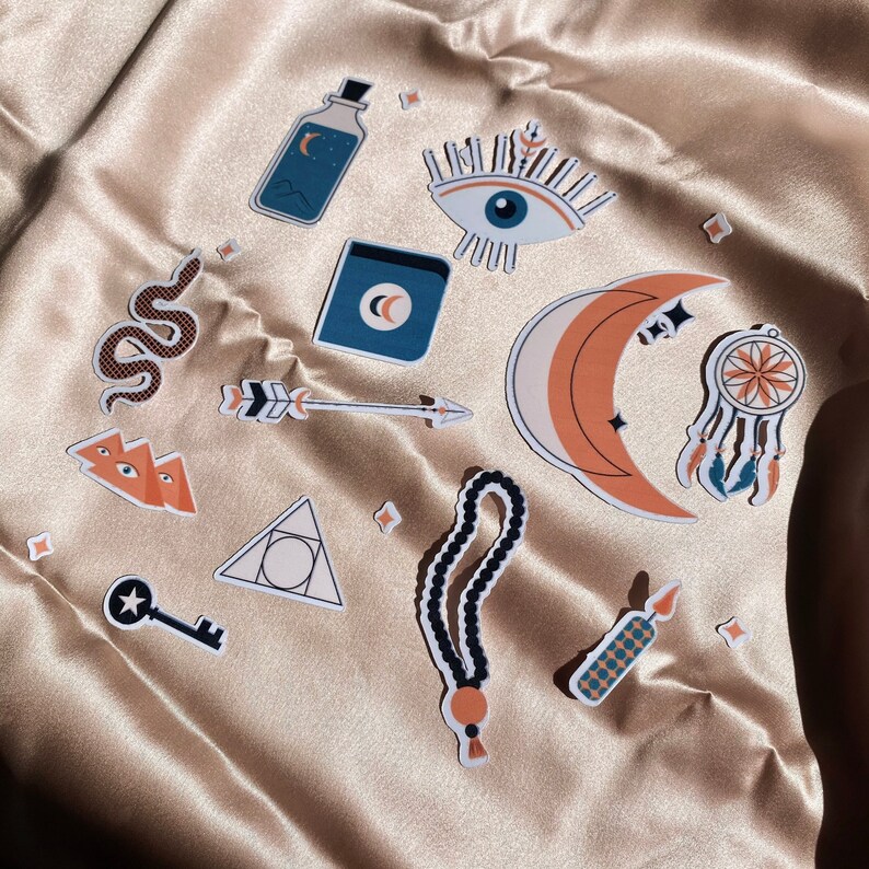 May include: A set of 10 colorful stickers featuring various designs, including a crescent moon, an eye, a snake, a key, a dreamcatcher, a bottle, an arrow, a triangle, a prayer beads, and a candle. The stickers are on a beige fabric background.